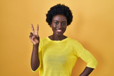 African young woman standing over yellow studio smiling looking to the camera showing fingers doing victory sign. number two. 