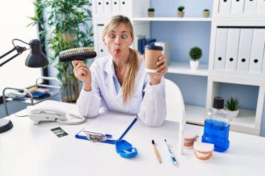 Beautiful dentist woman holding denture and toothbrush making fish face with mouth and squinting eyes, crazy and comical. 