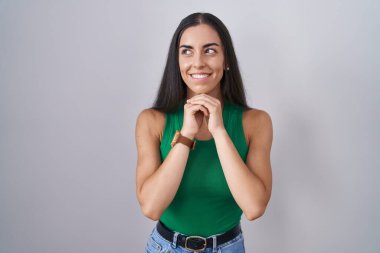 İzole edilmiş arka planda duran genç bir kadın gergin ve heyecanlı, elleri çenesinde yan tarafa bakıyor. 