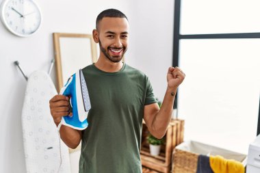 African american man holding electric iron screaming proud, celebrating victory and success very excited with raised arm 