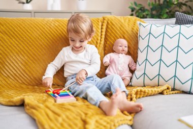 Ksilofon çalan sevimli beyaz kız evde oyuncak bebekle kanepede oturuyor.