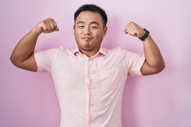 Chinese young man standing over pink background showing arms muscles smiling proud. fitness concept. 