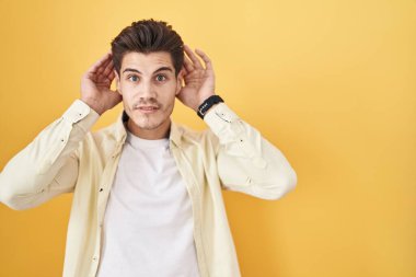 Sarı arka planda dikilen genç İspanyol adam iki elini de kulak hareketlerinde, dedikoduya meraklı olarak duymaya çalışıyor. İşitme sorunu, sağır 