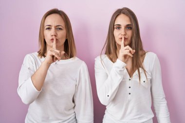 Middle age mother and young daughter standing over pink background asking to be quiet with finger on lips. silence and secret concept. 