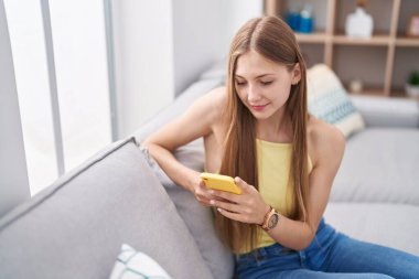 Beyaz kadın akıllı telefon kullanıyor. Evdeki kanepede oturuyor.