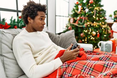 Genç Afrikalı Amerikalı adam evdeki Noel ağacının yanında kanepede oturmuş video oyunu oynuyor..
