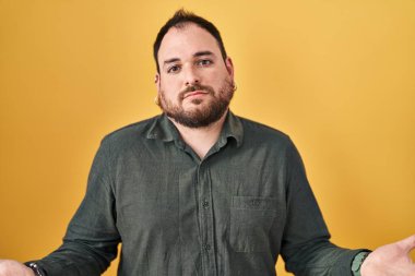 Plus size hispanic man with beard standing over yellow background clueless and confused with open arms, no idea concept. 