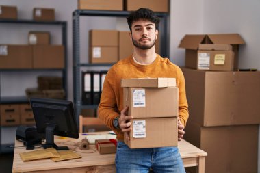 Hispanic man with beard working at small business ecommerce holding packages relaxed with serious expression on face. simple and natural looking at the camera. 