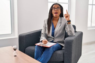 Young african american with braids working at consultation office amazed and surprised looking up and pointing with fingers and raised arms. 