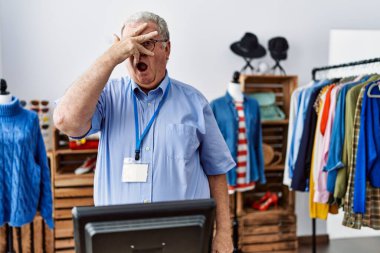 Senior man with grey hair working as manager at retail boutique peeking in shock covering face and eyes with hand, looking through fingers with embarrassed expression. 