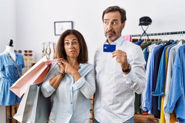 Hispanic middle age couple holding shopping bags and credit card pointing aside worried and nervous with forefinger, concerned and surprised expression 