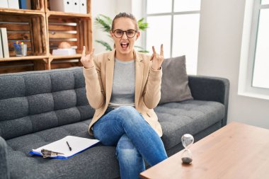 Young woman working at consultation office shouting with crazy expression doing rock symbol with hands up. music star. heavy music concept. 