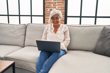 Orta yaşlı bir kadın evdeki koltukta dizüstü bilgisayar kullanıyor.
