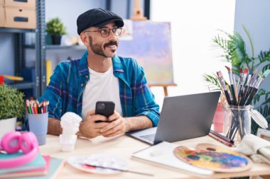 Sanat stüdyosunda dizüstü bilgisayar ve akıllı telefon kullanan genç bir İspanyol sanatçı.