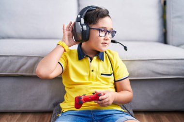 Young hispanic kid playing video game holding controller wearing headphones smiling with hand over ear listening an hearing to rumor or gossip. deafness concept. 