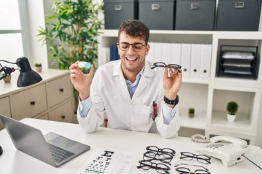 Young hispanic oculist holding glasses and contact lenses smiling and laughing hard out loud because funny crazy joke. 
