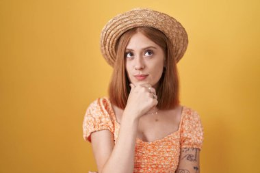 Young redhead woman standing over yellow background wearing summer hat looking confident at the camera with smile with crossed arms and hand raised on chin. thinking positive. 