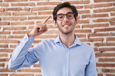 Young hispanic man standing over brick wall background smiling pointing to head with one finger, great idea or thought, good memory 