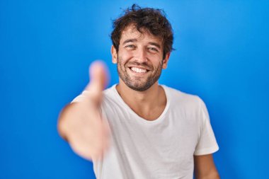 Mavi arka planda duran İspanyol genç adam dostça bir karşılama ve selamlama olarak el sıkışmayı sunuyor. başarılı iş. 