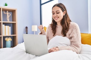Young hispanic woman using computer laptop on the bed pointing thumb up to the side smiling happy with open mouth 
