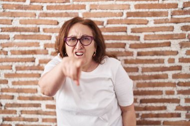 Senior woman with glasses standing over bricks wall pointing displeased and frustrated to the camera, angry and furious with you 