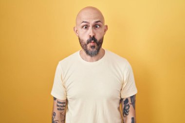 Hispanic man with tattoos standing over yellow background making fish face with lips, crazy and comical gesture. funny expression. 