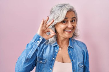 Middle age woman with grey hair standing over pink background smiling positive doing ok sign with hand and fingers. successful expression. 