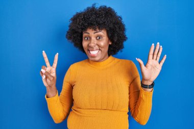 Black woman with curly hair standing over blue background showing and pointing up with fingers number seven while smiling confident and happy. 