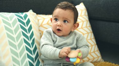 Adorable hispanic baby holding toy sitting on sofa at home