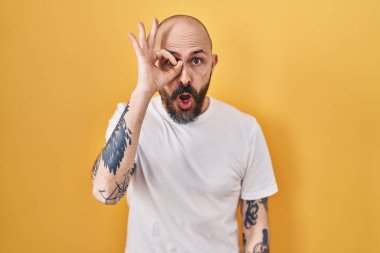Young hispanic man with tattoos standing over yellow background doing ok gesture shocked with surprised face, eye looking through fingers. unbelieving expression. 