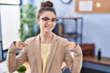 Teenager girl working at the office wearing glasses looking confident with smile on face, pointing oneself with fingers proud and happy. 