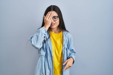 Genç İspanyol kadın mavi arka planda duruyor. Yarı yüzünü, göz ve ağzını eliyle kapatmaktan yorulmuş. Yüzüm acıyor.. 