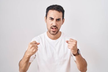 Handsome hispanic man standing over white background angry and mad raising fists frustrated and furious while shouting with anger. rage and aggressive concept. 