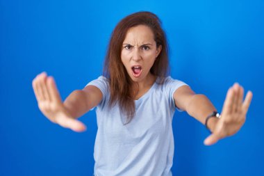 Brunette woman standing over blue background doing stop gesture with hands palms, angry and frustration expression 