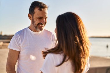 Orta yaşlı erkek ve kadın çift deniz kenarında yan yana duruyorlar.
