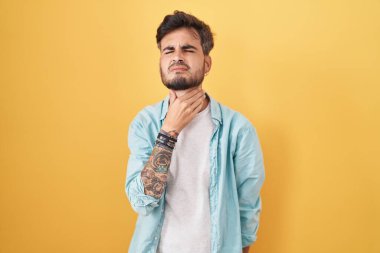 Young hispanic man with tattoos standing over yellow background touching painful neck, sore throat for flu, clod and infection 
