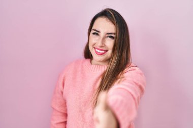 Young hispanic woman standing over pink background smiling friendly offering handshake as greeting and welcoming. successful business. 