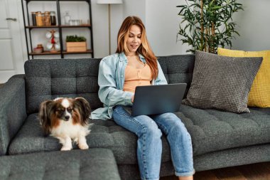 Genç beyaz kadın dizüstü bilgisayar kullanıyor. Kanepede köpeğiyle oturuyor.