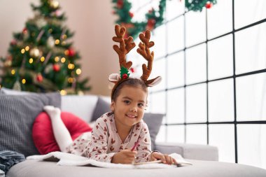 Adorable hispanic girl smiling confident lying on sofa by christmas tree at home