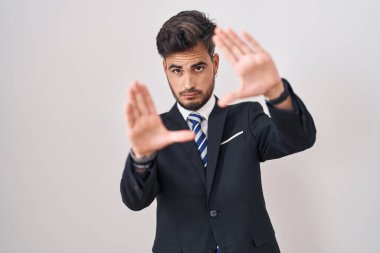 Young hispanic man with tattoos wearing business suit and tie doing frame using hands palms and fingers, camera perspective 