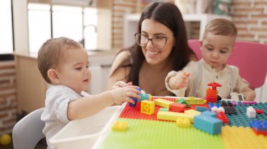 Öğretmen ve anaokulu öğrencileri anaokulundaki masada oturan inşaat bloklarıyla oynuyorlar.