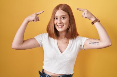 Young redhead woman standing over yellow background smiling pointing to head with both hands finger, great idea or thought, good memory 