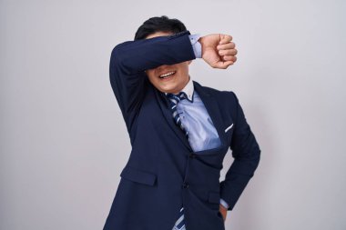 Young asian man wearing business suit and tie covering eyes with arm smiling cheerful and funny. blind concept. 
