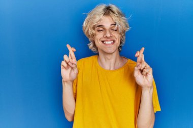Young modern man standing over blue background gesturing finger crossed smiling with hope and eyes closed. luck and superstitious concept. 