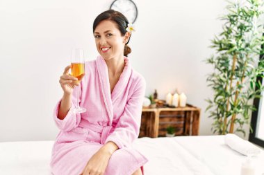Middle age hispanic woman toasting with champagne sitting on massage table at beauty center