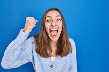 Mavi arka planda öfkeli ve kızgın duran genç bir kadın öfkeyle bağırırken yumruğunu sallıyor ve öfkeleniyor. öfke ve agresif konsept. 