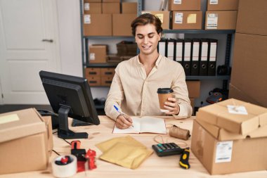 Young caucasian man ecommerce business worker writing on notebook drinking coffee at office