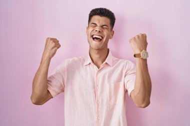 Young hispanic man standing over pink background celebrating surprised and amazed for success with arms raised and eyes closed. winner concept. 