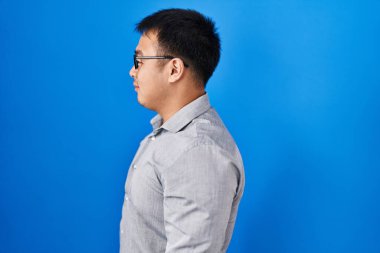 Young chinese man standing over blue background looking to side, relax profile pose with natural face with confident smile. 