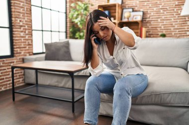 Young beautiful hispanic woman talking on smartphone with worried expression at home